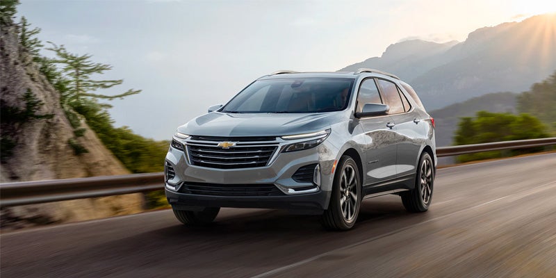 Light gray Chevrolet Equinox SUV driving on a winding road with mountains and a bright sunburst in the background.
