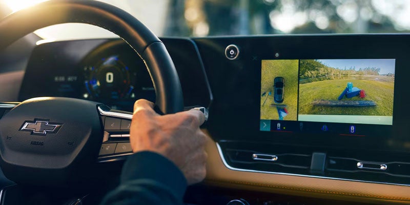 Hand on steering wheel in a car showing a multi-view camera display with aerial and rear views on the infotainment screen.