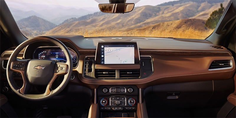 View from car's brown leather interior showing steering wheel, digital dash, infotainment screen with map, and mountains outside.