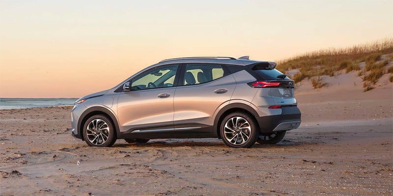 Silver Chevrolet Bolt EUV parked on a sandy beach with the ocean and dunes in the background at sunset.