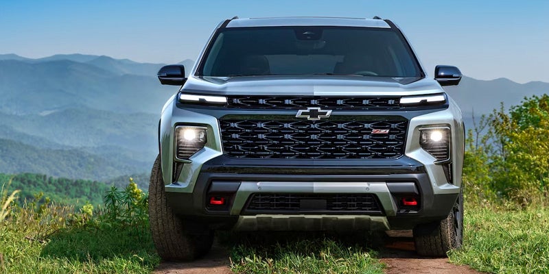 Silver Chevrolet SUV with a black grille and bright headlights on a dirt road, mountains in the background.