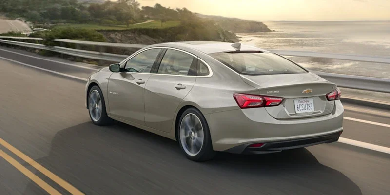 Light-colored sedan drives along a coastal road with the ocean in the background.