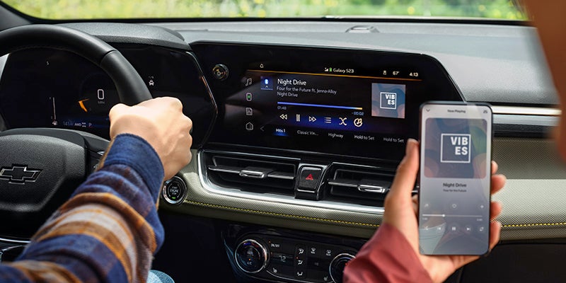 Driver's hands on steering wheel and phone, displaying synced 'Night Drive' music on the car's infotainment screen and phone.