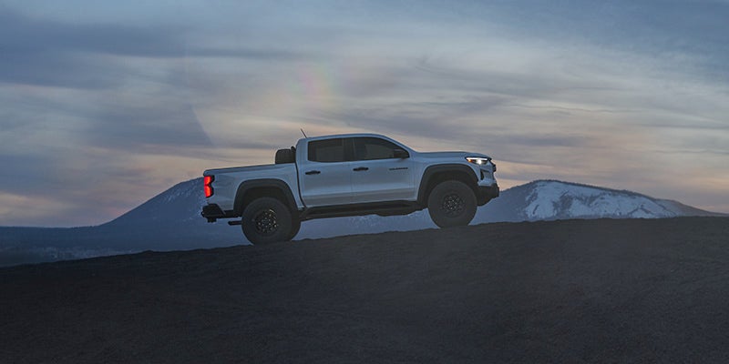 Light-colored pickup truck with illuminated headlights and taillights parked on a dark, rocky hill at dusk.