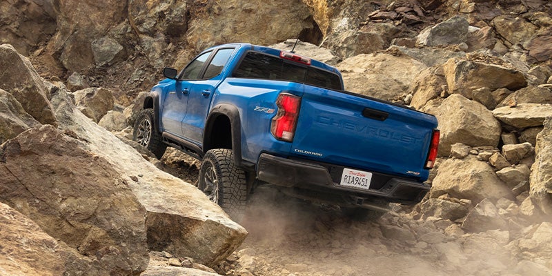 Blue Chevrolet Colorado pickup truck off-roading up a steep, rocky incline, kicking up dust.