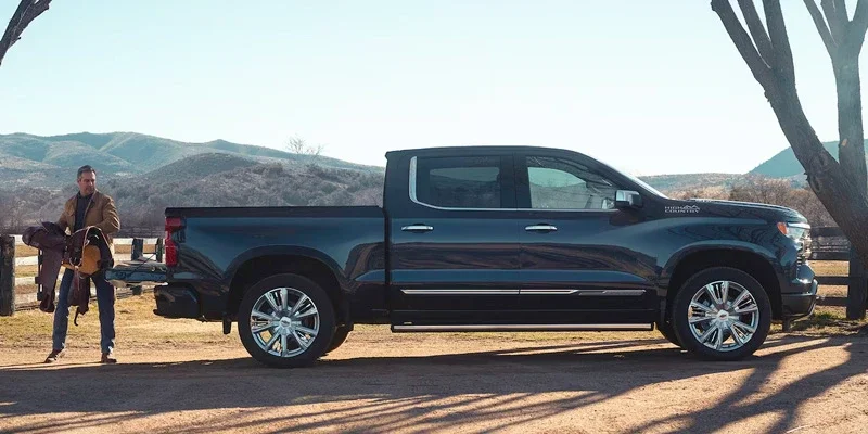 Man loads a saddle into the open tailgate of a dark pickup truck, with mountains in the background.
