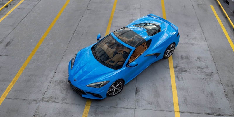 Blue sports car with a glass roof and brown interior, parked on gray asphalt with yellow lines, viewed from above.