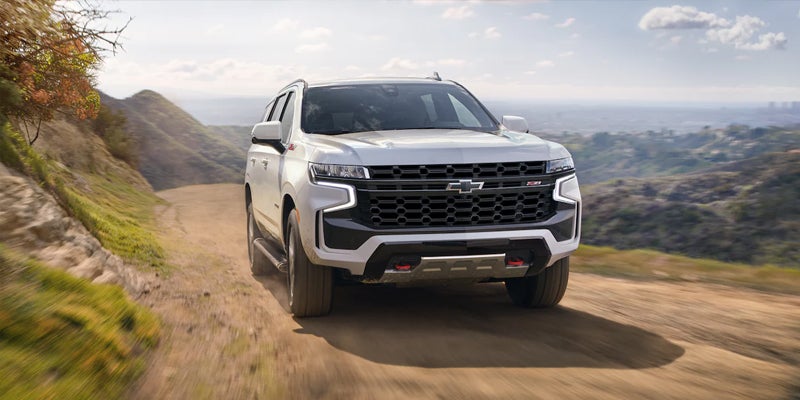 White Chevrolet Tahoe SUV driving on a dirt road, with mountains and a distant city visible under a partly cloudy sky.