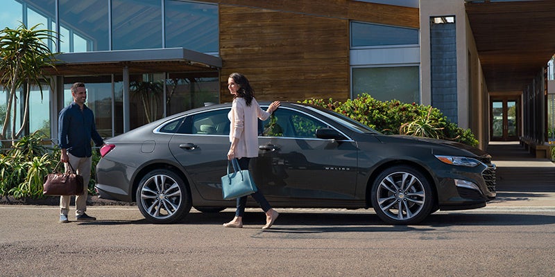 A man and woman approach a dark gray car parked in front of a modern house; she carries a blue bag, he a brown bag.