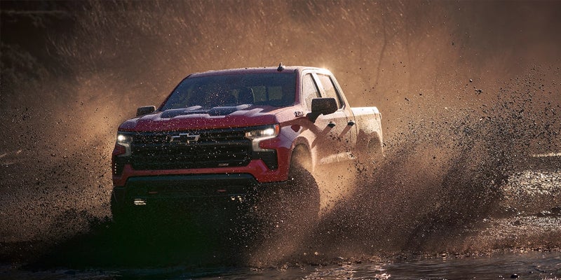 Red Chevrolet Silverado pickup truck splashes through mud and water, illuminated by sunlight.