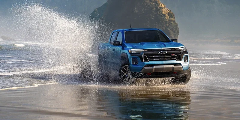 Blue Chevrolet Colorado pickup truck splashing through water on a beach with a rocky coastline in the background.