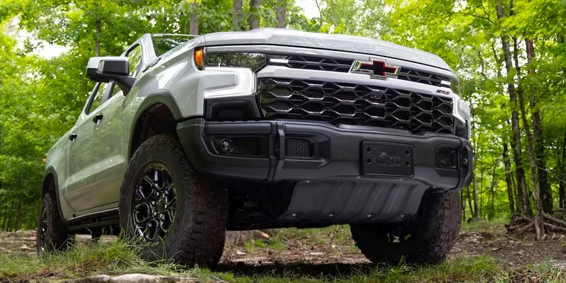 Silver Chevrolet pickup truck with large off-road tires, seen from a low front angle on a dirt trail in a forest.