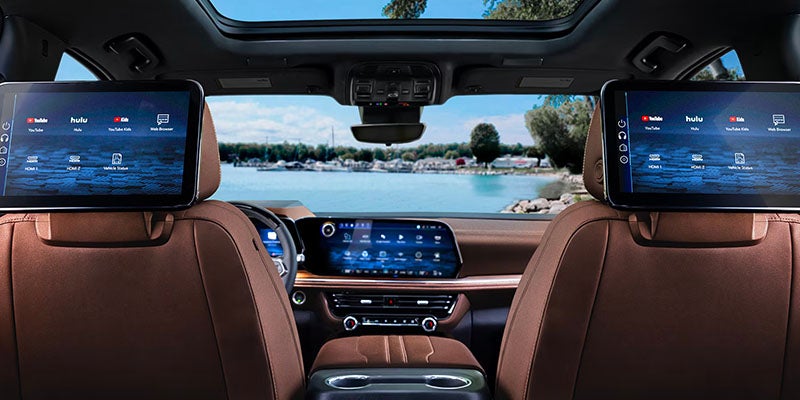 Car interior with two headrest screens showing entertainment apps, brown leather seats, and a lake view outside.