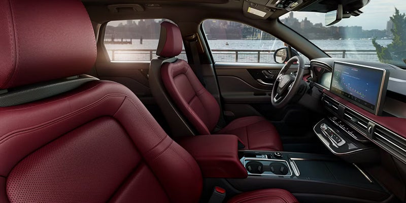 The luxurious car interior features red leather seats, dashboard, infotainment screen, and a view of a city waterfront through the windows.