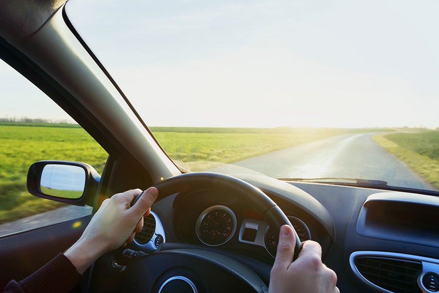 Hands on a steering wheel, driving on a winding road through green fields with bright sun ahead.