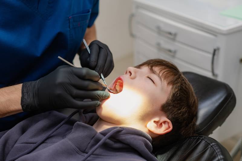 A dentist in black gloves and blue scrubs examines a child's mouth with dental tools.