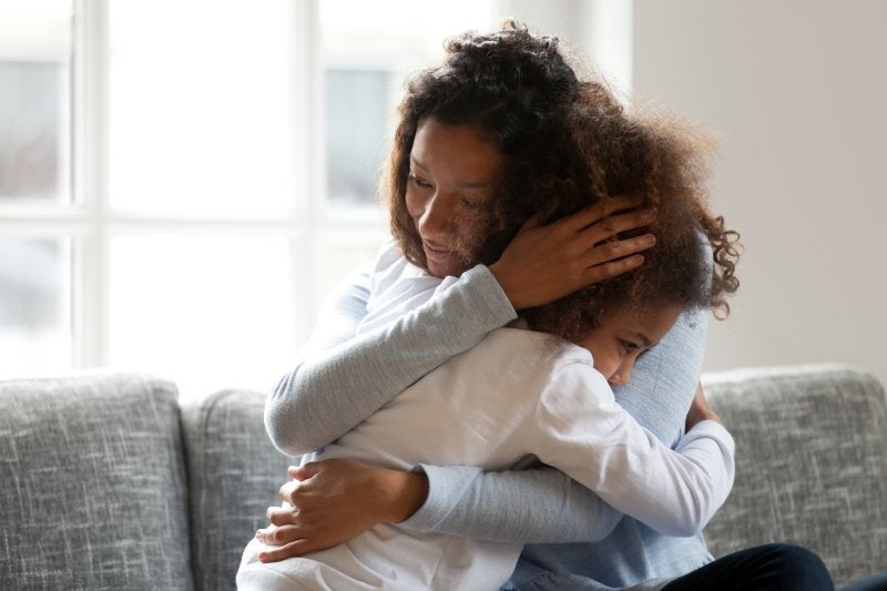 A caring mother with curly hair embraces her young daughter, who hides her face, on a gray couch.