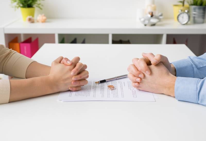 Clasped hands of two people on opposite sides of a table with a divorce document, pen, and wedding rings.