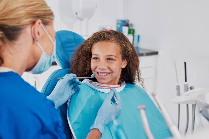A young girl with curly hair smiles while a gloved dentist examines her teeth with a mirror.