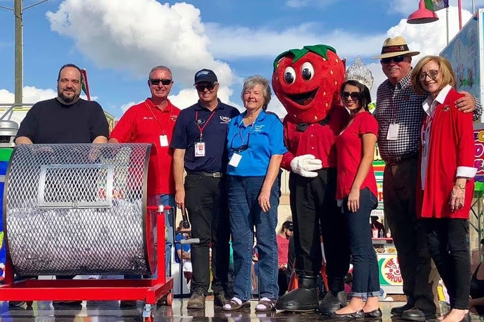 Group of 8 people, including a strawberry mascot, standing with a large raffle drum at an outdoor event.