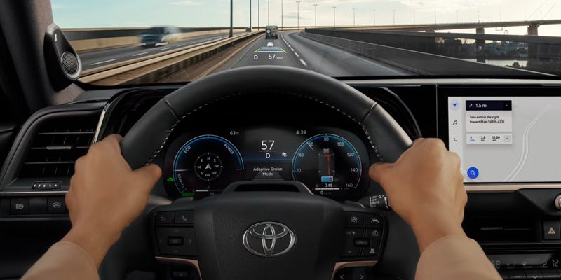 Interior view of a Toyota vehicle showing the driver’s hands on the steering wheel, digital instrument cluster, heads-up display, and navigation screen while driving on a highway.
