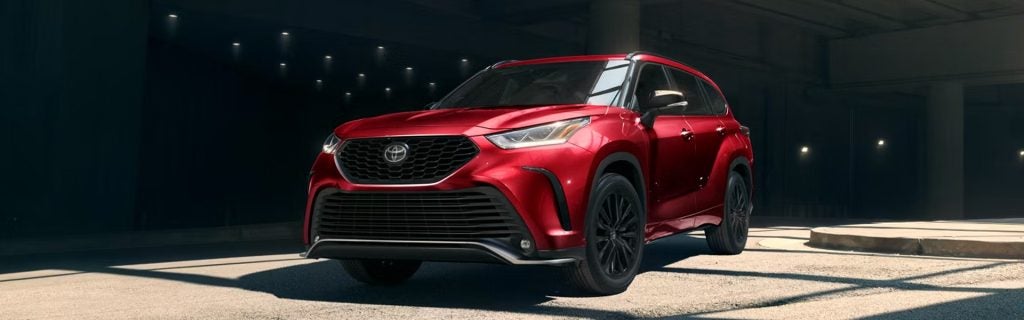 Red Toyota Highlander SUV parked on a sunlit concrete surface inside a modern, dimly lit architectural space with columns and overhead lights, shown in a front three-quarter view.