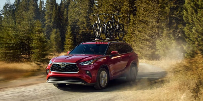 A red Toyota Highlander SUV with bicycles on the roof rack driving on a dirt road surrounded by trees and foliage.