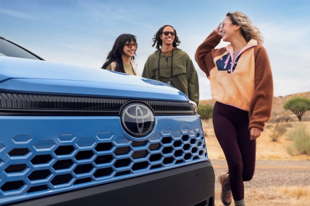 A blue Toyota vehicle in the foreground with three people in casual attire smiling and chatting in the background against a desert landscape.