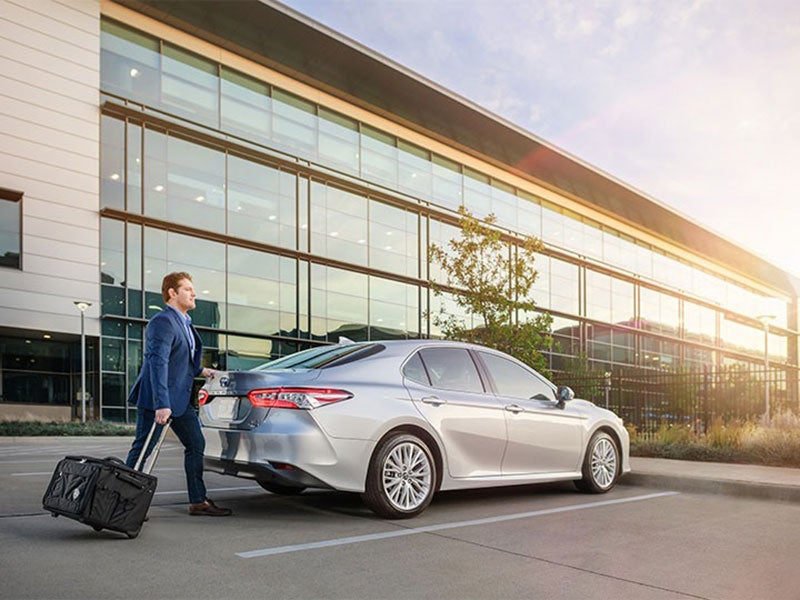 A businessman walks toward a silver sedan outside a modern office building with rolling luggage
