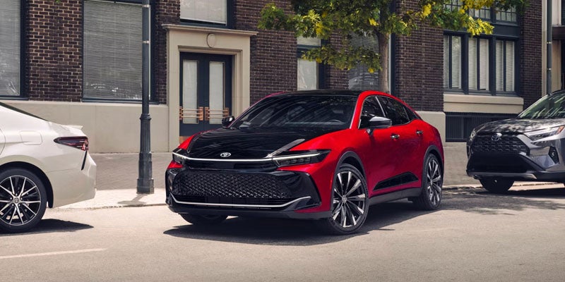 A red and black Toyota sedan parked on a street next to a white sedan and a dark SUV, with brick buildings in the background.