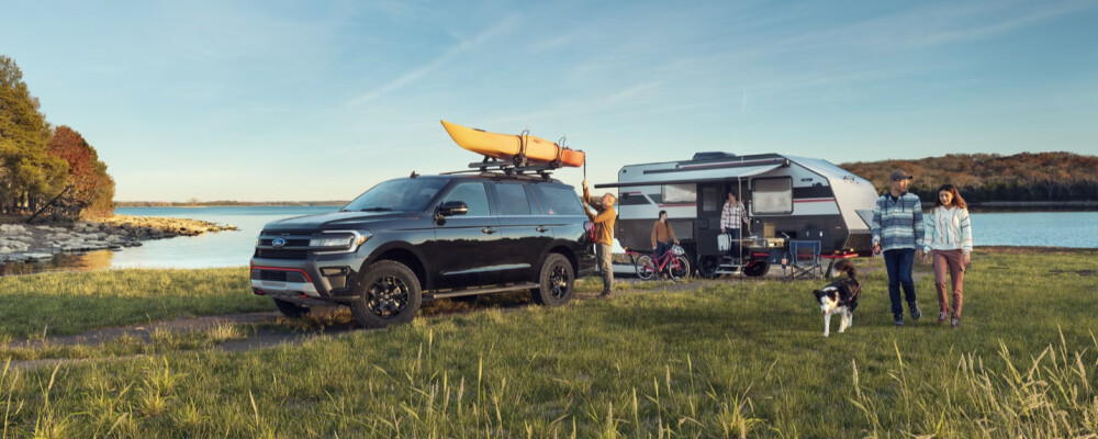 SUV with kayaks parked by a lake, family setting up beside a camper. A couple walks with a dog. Scenic, peaceful outdoor camping scene.
