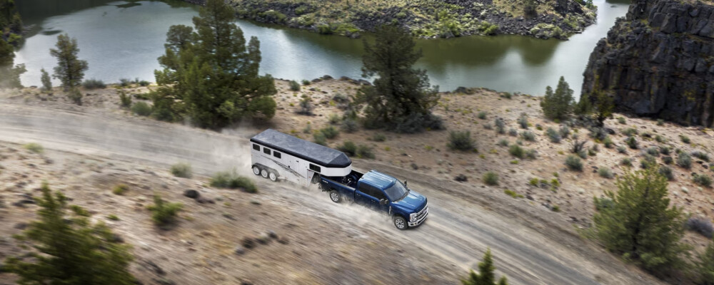 A blue pickup truck towing a large trailer speeds down a dirt road beside a scenic river, surrounded by trees and rocky cliffs. The scene conveys adventure.