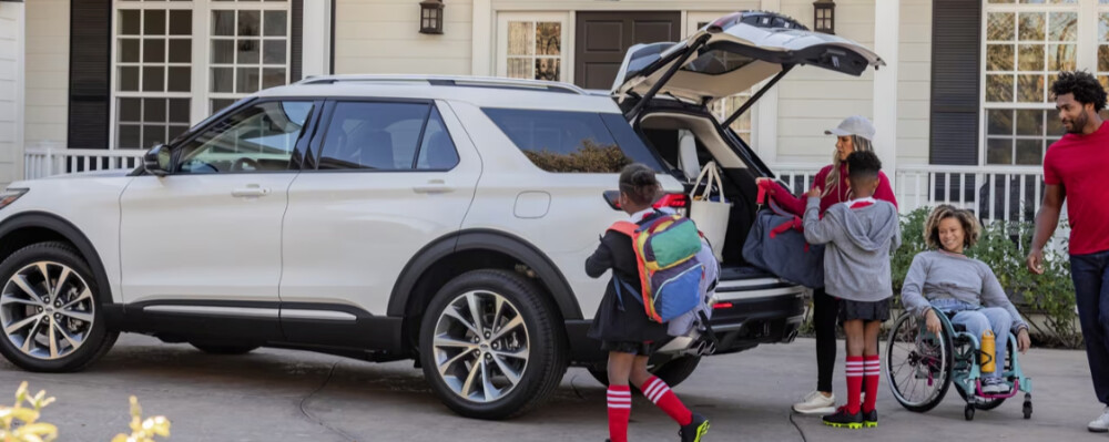 A family loads backpacks into a white SUV parked in a driveway. One child in a wheelchair smiles, conveying a happy, inclusive atmosphere.