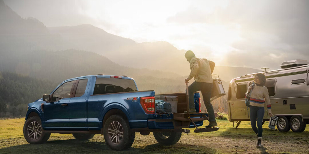 A blue pickup truck parked on a grassy field next to a silver trailer. A person steps off the truck bed, while another carries a lantern, with mountains in the background.