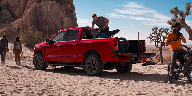 2025 Ford F-150 Lightning in red parked on desert terrain with tailgate down, loaded with electric bikes, as outdoor adventurers explore the rocky landscape in McMinnville, OR