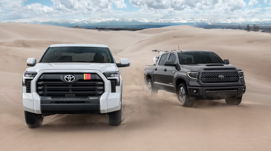 Side-by-side view of a white 2026 Toyota Tundra full-size truck and a dark grey Toyota pickup carrying a dirt bike driving through sand dunes, illustrating 2026 Toyota truck off-road utility and performance for Lake County drivers near Waukegan and Libertyville, IL.