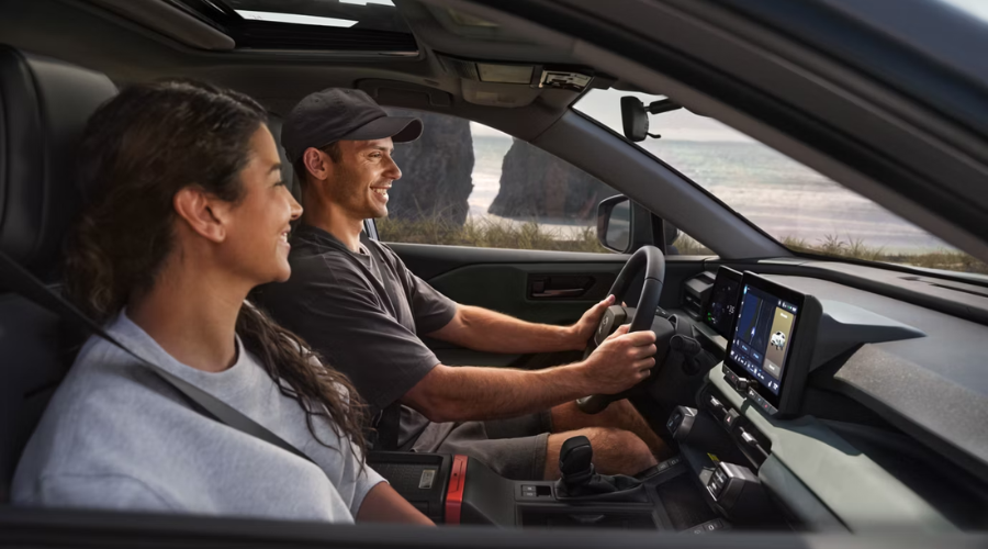 A smiling driver and passenger enjoying a scenic drive along the Lake Michigan coastline near Waukegan, IL in the 2026 Toyota RAV4, featuring the 10.5-inch Toyota Audio Multimedia touchscreen with Proactive Driving Assist and navigation display.