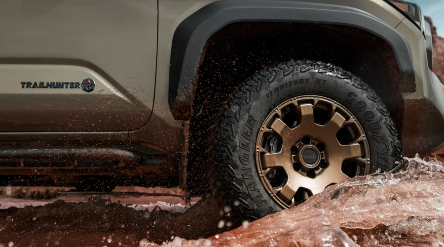 Close-up of a bronze wheel and Goodyear Wrangler all-terrain tire on a 2026 Toyota Tacoma Trailhunter splashing through deep mud and standing water, illustrating 4WD rain safety and wet road traction near Waukegan, IL.