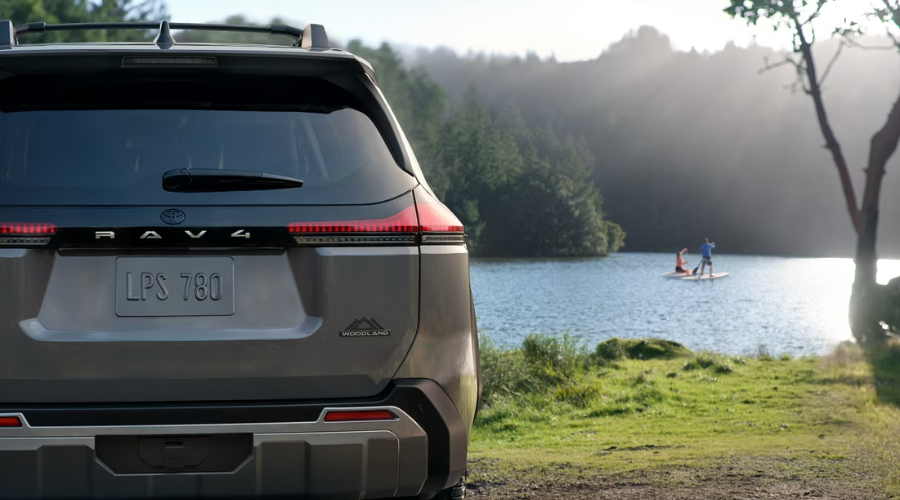 Rear view of a 2026 Toyota RAV4 Hybrid Woodland Edition showing the custom mountain badge, illuminated LED taillights, and roof rack parked beside a scenic lake near Antioch, IL while paddleboarders enjoy the water.