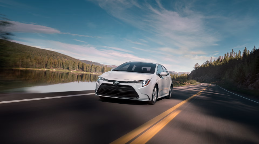 White 2027 Toyota Corolla LE sedan driving smoothly along a scenic highway next to a lake and forest landscape in Illinois available at Classic Toyota.