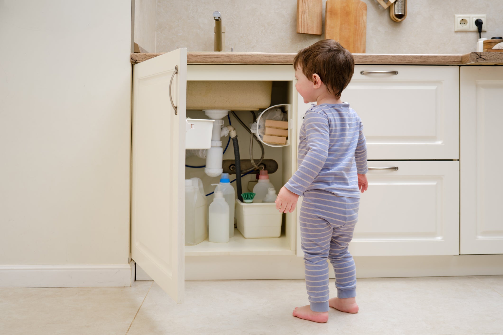 A child is playing with chemical cleaning products under the sink in the kitchen. Baby holds bottles with detergent. Kid aged about two years (one year nine months)