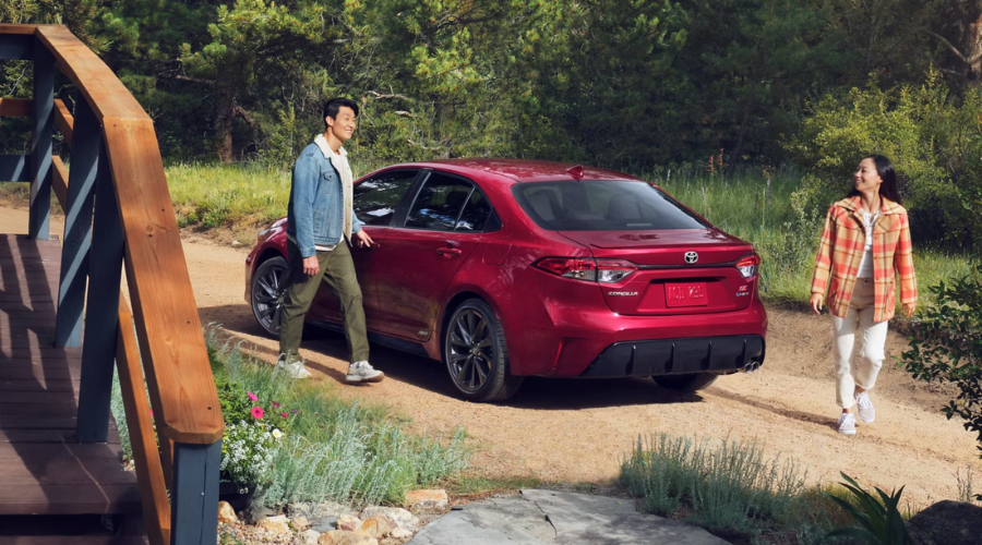 A couple walking toward a red 2026 Toyota Corolla Hybrid SE AWD parked on a rustic path in Lake County, IL, showcasing its outdoor versatility and efficiency for Waukegan and Gurnee commuters.