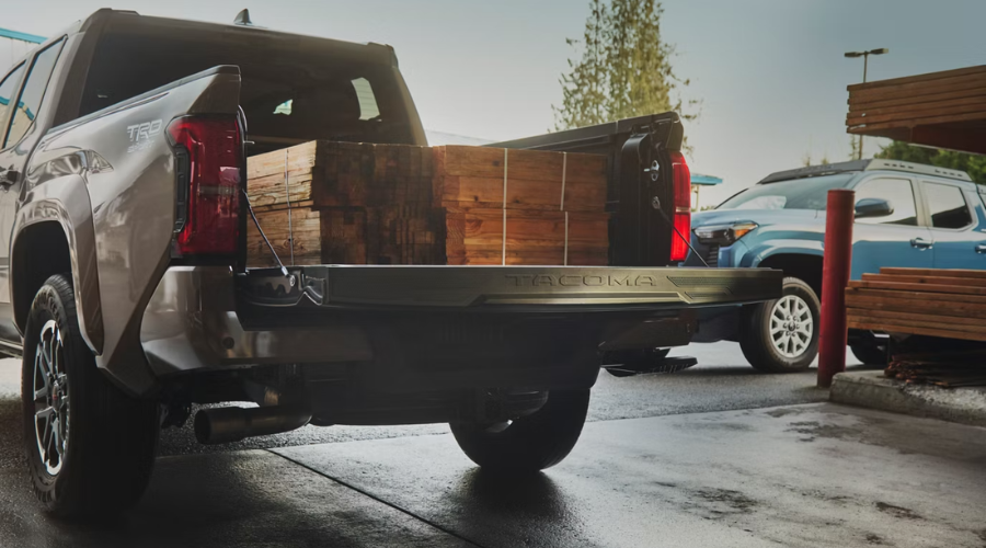 Rear three-quarter view of a bronze 2026 Toyota Tacoma TRD with the tailgate lowered, loaded with a heavy payload of stacked and strapped wooden lumber in Waukegan, Illinois 