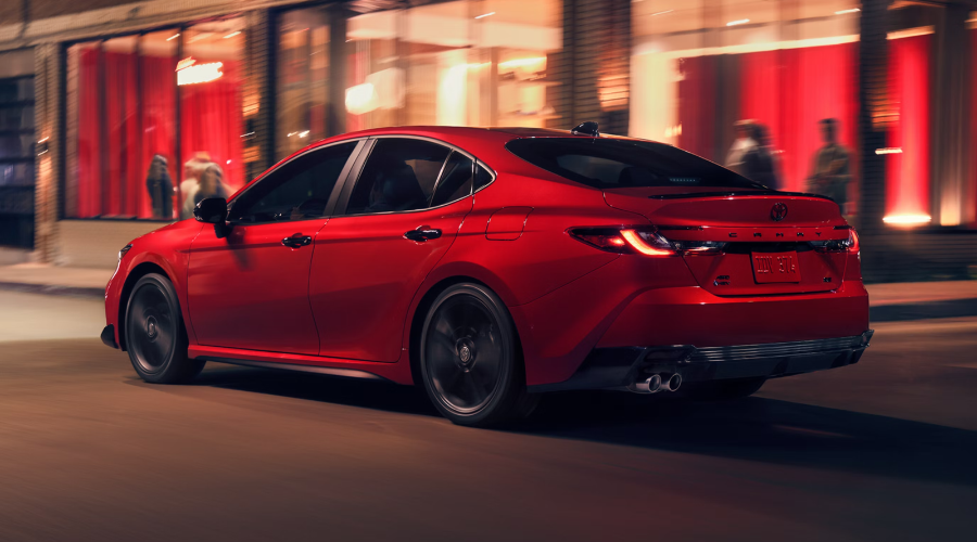 Side rear three-quarter view of a red 2026 Toyota Camry driving along a city street at night, showing rear AWD badging, illuminated LED taillights, and dual exhaust tips for Waukegan IL commuters at Classic Toyota.