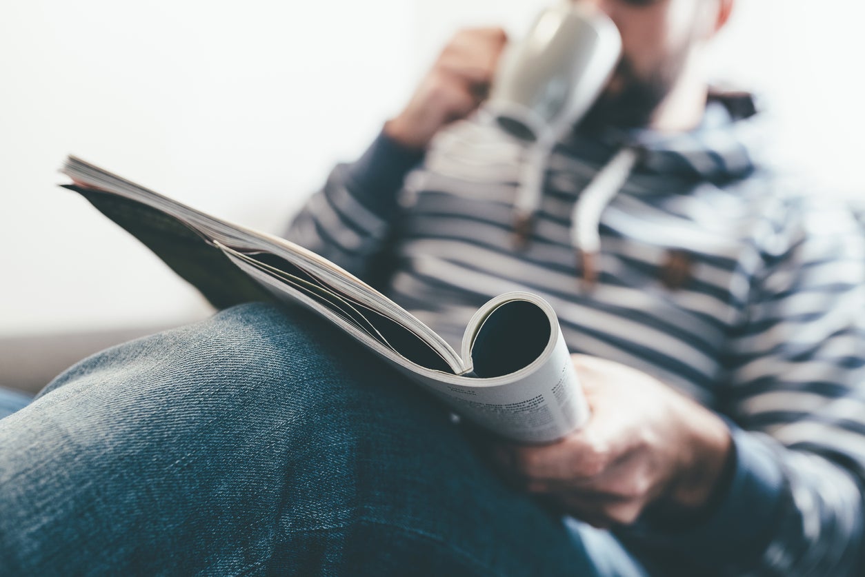 man reading magazine or newspaper and drinking coffee while relaxing on sofa