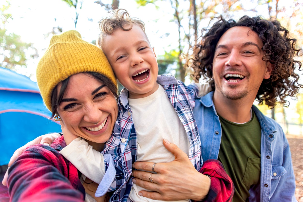 young happy family portrait camping outdoor with tent in autumn nature. mom, dad and little child making a pov selfie into the wild. people having fun together hugging each other smiling. lifestyle