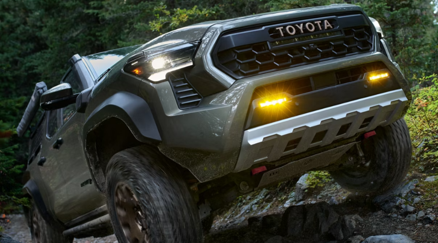 Low-angle front view of a dark green 2026 Toyota Tacoma off-road truck climbing rocky trail terrain with illuminated LED headlights and bumper fog lights, showcasing midsize capability for Lake County drivers comparing Tacoma vs Tundra models near Waukegan, IL.