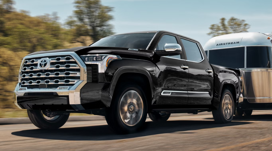 Black 2026 Toyota Tundra i-FORCE MAX full-size pickup truck with chrome grille towing a silver Airstream travel trailer along an open highway, demonstrating 12,000-pound towing capacity for Lake County drivers comparing Tacoma vs Tundra models near Libertyville and Waukegan, IL.