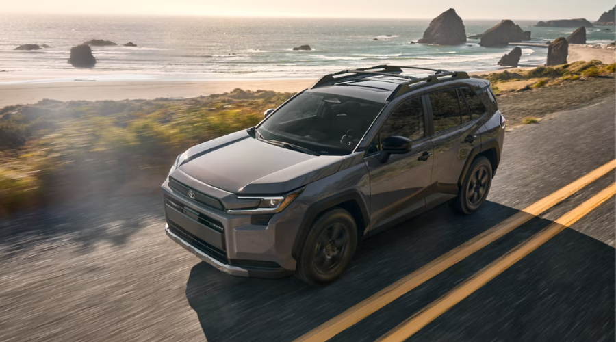 Exterior front-three-quarter view of a dark metallic 2026 Toyota RAV4 driving along a scenic coastal highway near Waukegan, IL, showcasing its front emblem millimeter-wave radar housing and rugged roof rack rails.