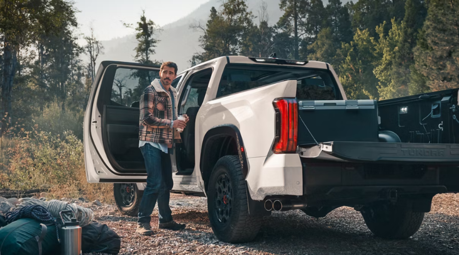 Man holding a coffee cup beside a white 2026 Toyota Tundra CrewMax with an open tailgate and loaded truck bed at an outdoor campsite, demonstrating weekend camping utility and bed storage for Lake County drivers near Waukegan and Libertyville, IL